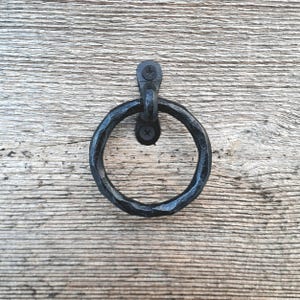 Op de afbeelding: Zwarte metalen ringtrekhengsel gemonteerd op een houten oppervlak. De hendel is gemaakt van gesmeed metaal en heeft een rustieke, gehammerde afwerking.