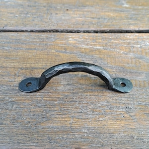 May include: Black metal drawer pull with a hammered finish. The pull has rounded ends and a slightly curved shape.