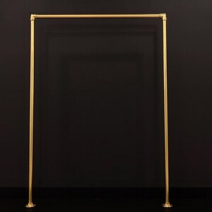 Peut inclure: Un portant à vêtements en métal doré avec un cadre rectangulaire. Il comporte une barre horizontale en haut et des supports verticaux. Chaque support a un pied circulaire. Le fond est noir.
