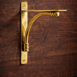 May include: A brass shelf bracket with a curved arm and a small, round detail at the end. The bracket is mounted on a dark brown wooden surface, showcasing its elegant design and finish. The bracket is designed to hold a shelf.