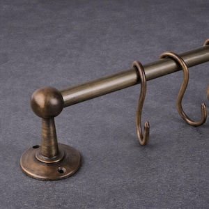 May include: Bronze-toned kitchen rail with three S-shaped hooks. The rail has a round ball finial on one end and a circular base for mounting. The hooks are evenly spaced along the rail, offering a classic, functional design.