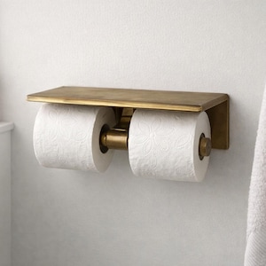 May include: A wall-mounted double toilet paper holder made of brass. The holder has a rectangular shelf above the two rolls of white toilet paper. A wooden toothbrush holder and a white towel are also visible.