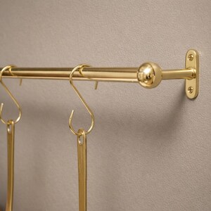May include: A brass kitchen pot rack with hanging S-hooks and copper pots. The rack is mounted on a neutral-colored wall. The design is simple and elegant, suitable for a classic kitchen.