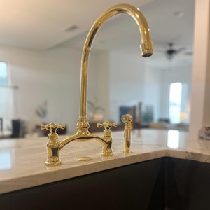 以下が含まれることがあります： 2つのハンドルと引き出し式のスプレイヤーが付いた金のキッチン蛇口。蛇口は白いカウンタートップに黒いシンクに取り付けられています。