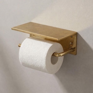 以下が含まれることがあります： 壁に取り付けられた金色の金属製トイレットペーパーホルダー。ホルダーにはロールの上に棚があり、紙を保持するための湾曲したバーがあります。白いトイレットペーパーのロールがセットされています。