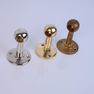 Op de afbeelding: Drie metalen stanghouders in zilver, goud en brons. Elk heeft een cirkelvormige basis met schroefgaten, een gebogen steel en een bolvormige bovenkant met een gat. De houders zijn gerangschikt op een wit oppervlak.