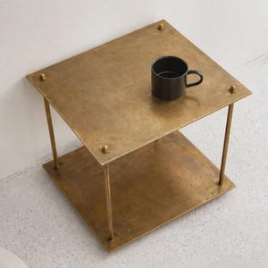 Può includere: Un tavolino quadrato a due livelli con finitura in ottone. Il tavolo ha un design semplice con un ripiano inferiore ed è sostenuto da quattro gambe sottili. Una tazza nera è appoggiata sulla superficie superiore. Il tavolo misura circa 46 cm quadrati.