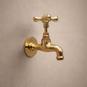 Könnte beinhalten: Ein an der Wand montierter Wasserhahn aus poliertem Messing. Der Wasserhahn hat einen Kreuzgriff und einen gebogenen Auslauf. Die runde Basis ist an einer neutralfarbenen Wand befestigt. Der Wasserhahn ist für den Wasserfluss konzipiert.