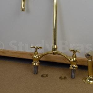 以下が含まれることがあります： 2つのハンドルと1つの注ぎ口を持つ金色のキッチン蛇口。蛇口は木製の表面に取り付けられており、クロムメッキのスプレースプレーヘッドが付いています。