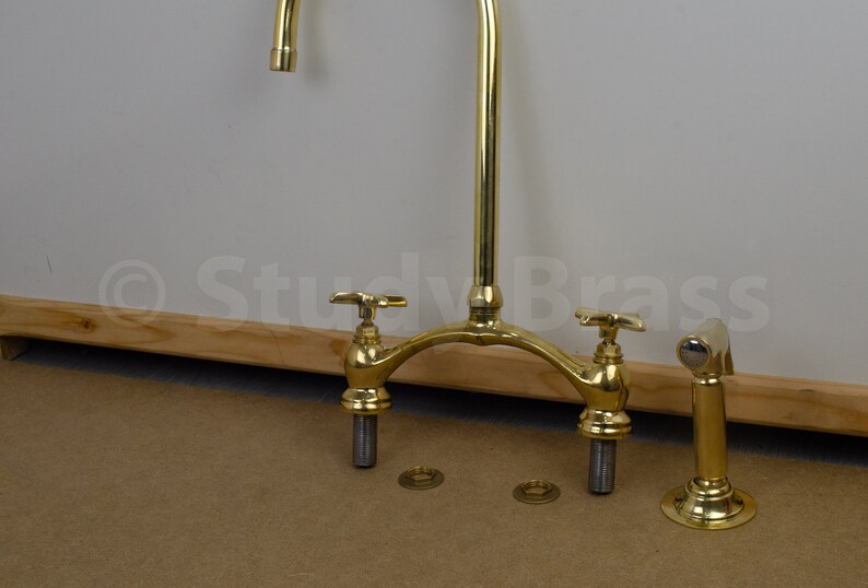 以下が含まれることがあります： 2つのハンドルと1つの注ぎ口を持つ金色のキッチン蛇口。蛇口は木製の表面に取り付けられており、クロムメッキのスプレースプレーヘッドが付いています。