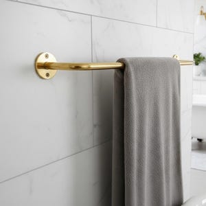 May include: A gold-colored towel bar mounted on a white tiled wall. A gray towel hangs from the bar. The bar has a circular mounting plate on one end and a curved design.