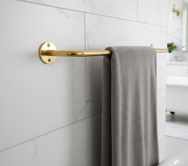 May include: A gold-colored towel bar mounted on a white tiled wall. A gray towel hangs from the bar. The bar has a circular mounting plate on one end and a curved design.