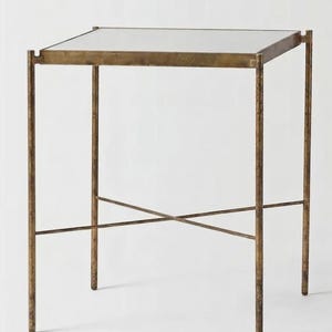 Puede incluir: Una mesa auxiliar cuadrada con una tapa de espejo y un marco de metal color bronce. La mesa tiene cuatro patas rectas y un soporte en forma de X. La mesa mide aproximadamente 61 cm de altura.
