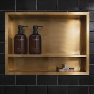 May include: A gold-toned, recessed shelf built into a black tiled wall. Two brown shampoo bottles with black pumps are on the top shelf. The word "SHAMPOO" is printed on the bottles. A small brush sits on the lower shelf.