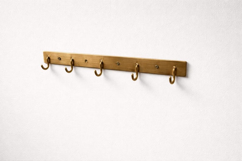 May include: A brass-toned wall-mounted coat rack with six curved hooks. The rack is rectangular with rounded edges and is mounted on a white wall. The hooks are evenly spaced along the bottom edge of the rack.