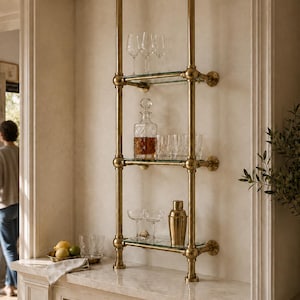 May include: A floor-to-ceiling shelving unit featuring two vertical brass pipes and three horizontal glass shelves. The unit is mounted to the wall and ceiling, displaying clear glassware, a glass decanter, and a brass cocktail shaker.