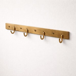 May include: A brass-coloured wall-mounted hook rack with five curved hooks. The rectangular board is mounted on a white wall. The hooks are evenly spaced along the board, providing a stylish storage solution.
