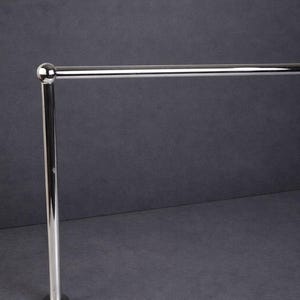 Puede incluir: Un perchero cromado con un diseño minimalista. El perchero presenta una barra horizontal sostenida por postes verticales, con un detalle de bola redondeada en la esquina. El metal pulido refleja la luz sobre un fondo gris oscuro.