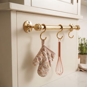 May include: A polished brass kitchen rail mounted horizontally with three matching S-hooks. A floral print oven mitt hangs from the first hook, and a copper-toned wire whisk hangs from the second hook, leaving the third hook empty.