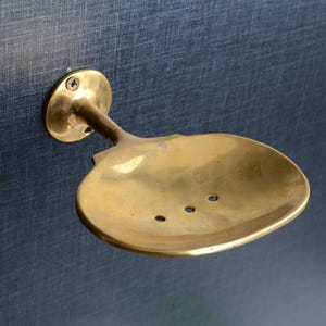 Op de afbeelding: Een messing zeepbakje met een lepelachtig ontwerp, gemonteerd op een donkerblauwe, getextureerde ondergrond. Het bakje heeft drie afvoergaten en een ronde montageplaat met een enkele schroef.