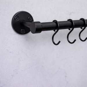 May include: A black metal kitchen rail with several S-shaped hooks. The rail is mounted on a wall with a circular bracket and screws. The design is minimalist and industrial, suitable for hanging kitchen utensils.