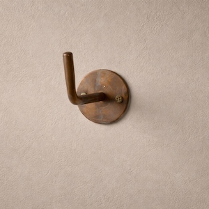 Peut inclure: Un crochet mural de couleur bronze avec une base circulaire et un crochet incurv&eacute;. Le crochet est mont&eacute; sur un mur textur&eacute; de couleur claire. Le crochet est fix&eacute; avec une seule vis.