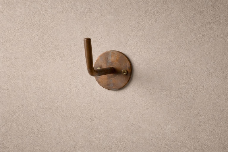 Peut inclure: Un crochet mural de couleur bronze avec une base circulaire et un crochet incurv&eacute;. Le crochet est mont&eacute; sur un mur textur&eacute; de couleur claire. Le crochet est fix&eacute; avec une seule vis.