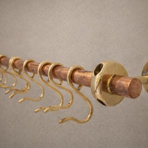 Könnte beinhalten: Eine Kupfer- und Messing-Hängestange mit acht goldfarbenen Haken. Die Stange ist an einer Wand befestigt und verfügt über eine Kupferstange mit Messingakzenten und kreisförmigen Montageplatten. Die Haken sind gleichmäßig verteilt.