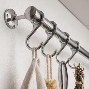 Puede incluir: Una barra de cocina de acero inoxidable con ganchos en forma de S, que sostiene una toalla beige, un cepillo, un utensilio y un manojo de hierbas. Montada en una pared blanca, ofrece almacenamiento.