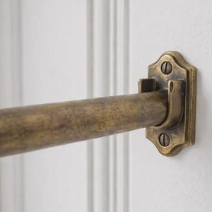 May include: Close-up of a brass-toned curtain rod and bracket mounted on a white wall. The rod is cylindrical, and the bracket has a decorative, ornate design with two screws. The finish has a vintage, aged appearance.