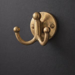 May include: A brass double hook with a circular backplate. The hook has a rounded top and two curved arms with ball-shaped ends. The fixture is mounted on a dark gray surface, showcasing its warm, metallic finish. Ideal for hanging items.