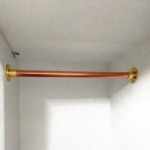 May include: A copper closet rod with brass mounting hardware. The rod is a warm, reddish-brown color and is mounted horizontally between two white walls. The brass hardware adds a touch of elegance to the functional design.