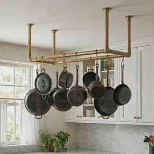 Może przedstawiać: Złoty, montowany do sufitu wieszak na garnki z kilkoma czarnymi garnkami i patelniami. Stojak wykonany jest z metalowych rur i znajduje się w kuchni z białymi szafkami i marmurowym blatem.