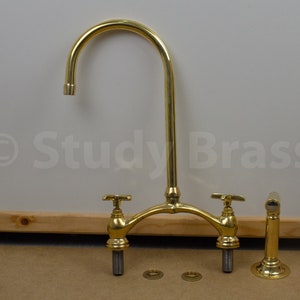 以下が含まれることがあります： 2つのハンドルと高い注ぎ口を持つ金色のキッチン蛇口。蛇口は分解されて木製の表面に置かれた部品と共に示されています。蛇口には「Study Brass」というラベルが貼られています。