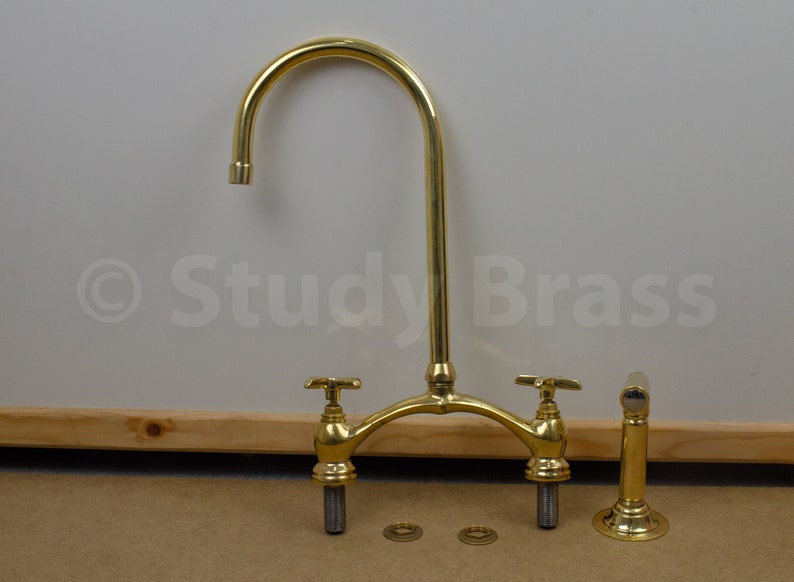 以下が含まれることがあります： 2つのハンドルと高い注ぎ口を持つ金色のキッチン蛇口。蛇口は分解されて木製の表面に置かれた部品と共に示されています。蛇口には「Study Brass」というラベルが貼られています。