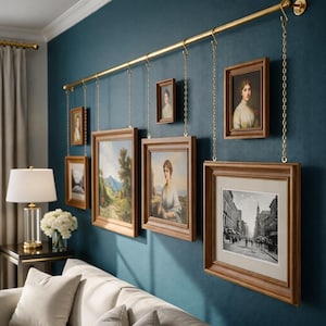 May include: A collection of framed art pieces hanging from a gold-colored rod on a teal wall. The art includes portraits and landscape paintings, each in a wooden frame. The frames are suspended by chains.