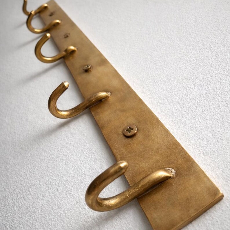 May include: A brass-toned wall-mounted hook rack with four curved hooks. The rectangular backplate is secured with screws. The hooks are evenly spaced and project outward, offering a simple, functional design for hanging items.