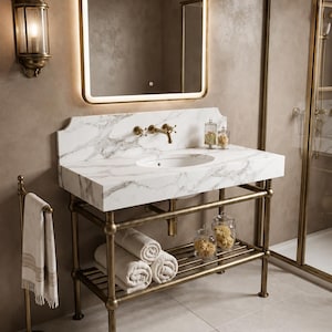Puede incluir: Un mueble de baño con encimera de mármol blanco con vetas grises, lavabo ovalado y grifo de latón. La base está hecha de tuberías de latón con un estante de listones que sostiene toallas blancas enrolladas y frascos de vidrio.