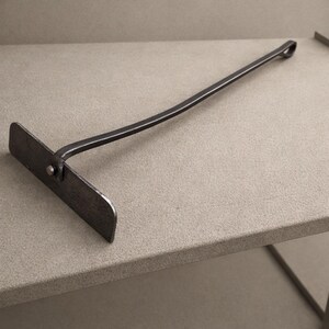 May include: A handcrafted, dark metal fire poker with a flat, rectangular head and a long, curved handle ending in a loop. The tool is designed for managing embers and logs in a fireplace or fire pit.