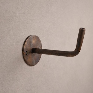 Peut inclure: Un crochet mural de couleur bronze avec une base circulaire et un bras incurv&eacute;. Le crochet est mont&eacute; sur un mur gris clair. Le crochet est en m&eacute;tal et a un aspect vintage ou antique.