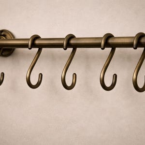 Könnte beinhalten: Eine bronzefarbene Küchenreling mit sechs gebogenen Haken. Die Reling ist an einer Wand befestigt und hat ein dekoratives Endstück. Die Haken sind gleichmäßig entlang der Reling angeordnet und zum Aufhängen von Küchenutensilien bestimmt.