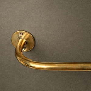 May include: A curved, brass-toned towel bar with a circular mounting plate. The bar is attached to a dark gray wall. The mounting plate is secured with two screws.