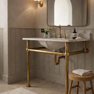 以下が含まれることがあります： 大理石の洗面台と真鍮製の金具を備えたバスルーム。洗面台は真鍮製のスタンドで支えられています。小さな籐のスツールと柔らかいラグが空間を完成させ、温かい雰囲気を作り出しています。