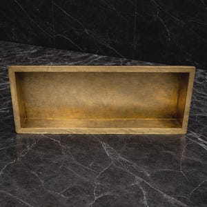 May include: A rectangular, open-top container with a brass finish. The container is set against a dark, marbled surface. The brass has a slightly aged appearance, with variations in tone. The overall design is simple and modern.