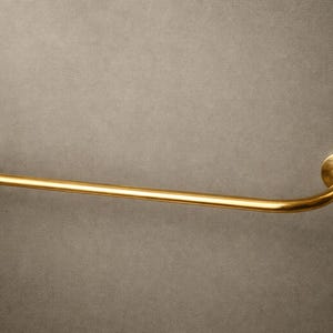 May include: A gold-colored towel bar with a curved design. The bar is mounted on a gray wall. The bar has a circular mounting plate on each end, with a curved section connecting them. The bar is made of metal.