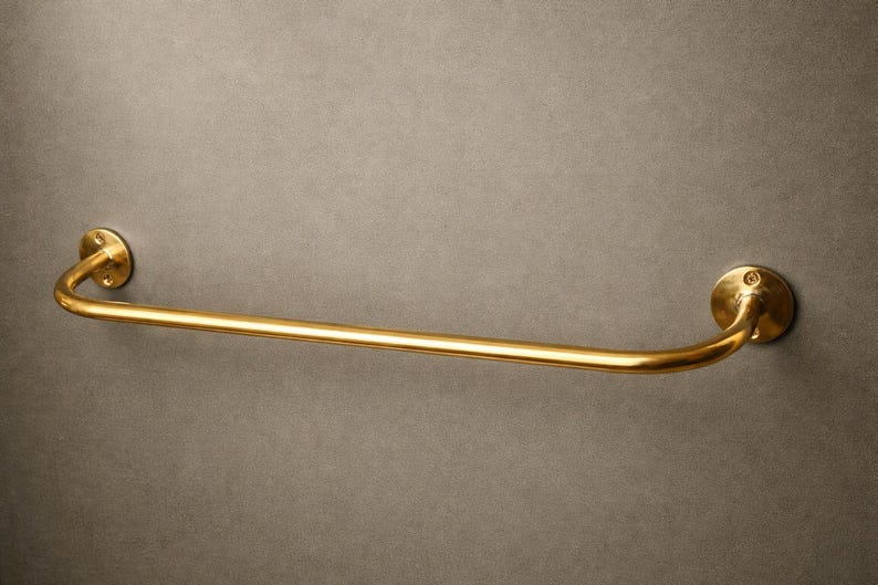 May include: A gold-colored towel bar with a curved design. The bar is mounted on a gray wall. The bar has a circular mounting plate on each end, with a curved section connecting them. The bar is made of metal.