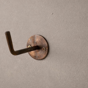 Peut inclure: Un crochet mural de couleur bronze avec une base circulaire et un bras incurv&eacute;. Le crochet est mont&eacute; sur un mur textur&eacute; gris clair. La base est fix&eacute;e avec deux vis.