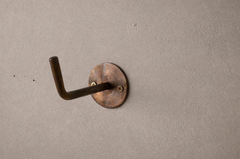 Peut inclure: Un crochet mural de couleur bronze avec une base circulaire et un bras incurv&eacute;. Le crochet est mont&eacute; sur un mur textur&eacute; gris clair. La base est fix&eacute;e avec deux vis.