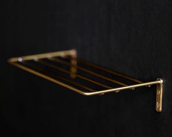 May include: A wall-mounted, gold-colored metal shelf with a rectangular design. The shelf has multiple horizontal bars and is attached to the wall with two brackets. The background is a solid black color.