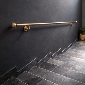 Puede incluir: Una barandilla de latón con un extremo redondeado está montada en una pared gris oscuro. La barandilla corre horizontalmente sobre una escalera de piedra gris. Se ve una pequeña planta en una maceta tejida.